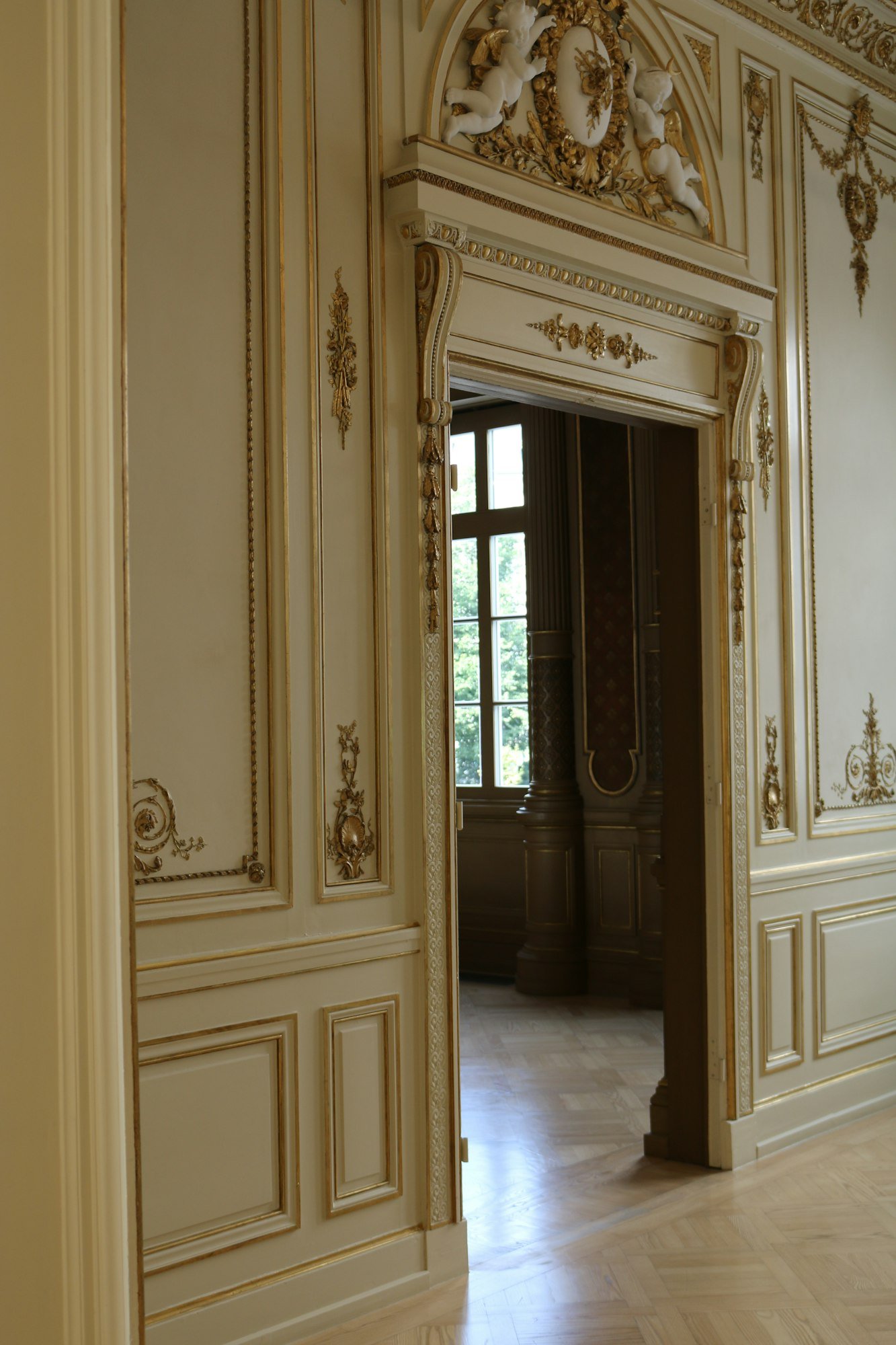 Des boiseries et moulures dorées autour d’une porte double, sur un parquet en chevrons